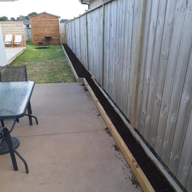 The finished bed from the patio, the timber edge and dark soil running the length of the fence with the shed and lawn beyond