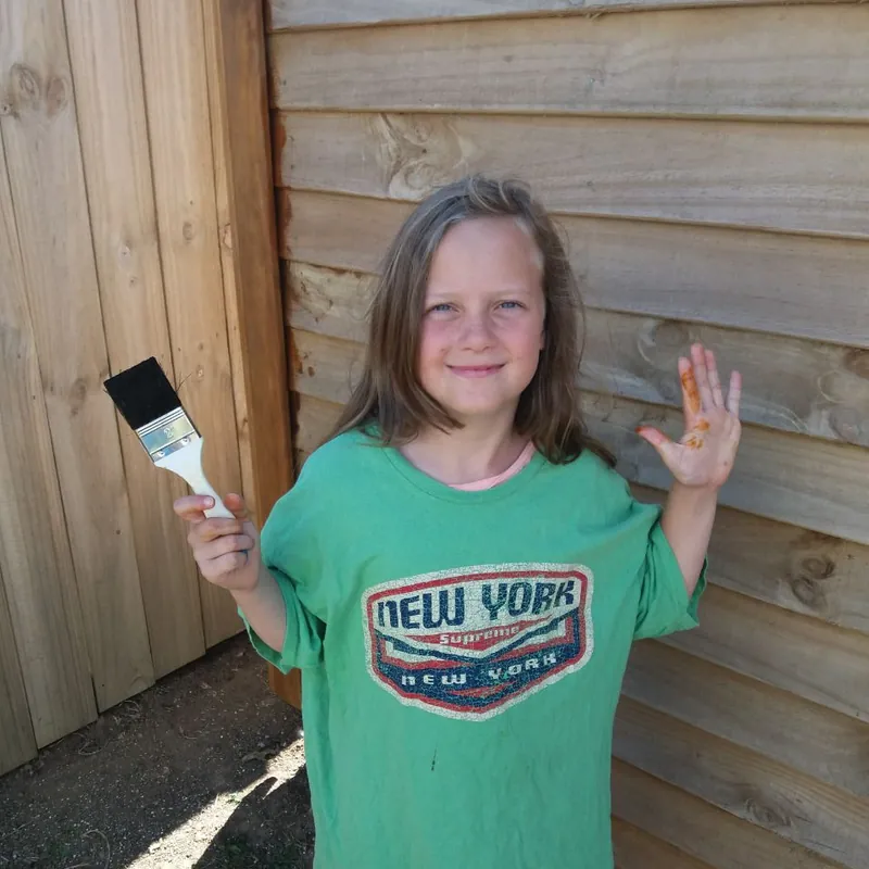 One of the girls at the shed in 2021, holding up a paintbrush and a hand covered in stain