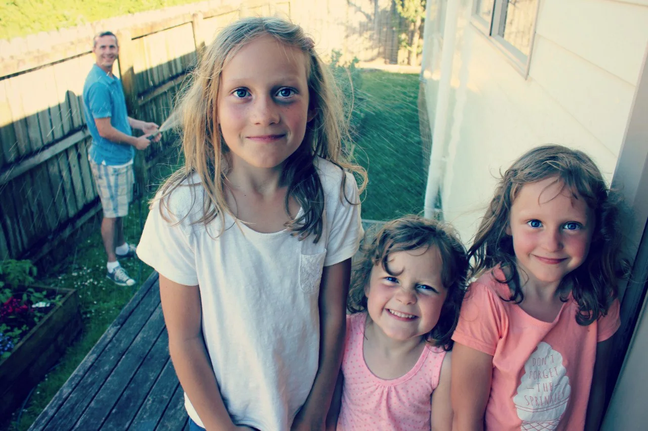 The three girls lined up on the deck, soaked through and grinning at the camera, with me in the background at the fence holding the hose