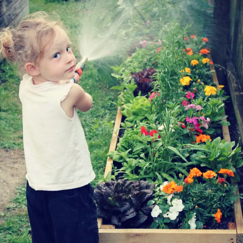 One of the girls with the hose over her shoulder beside a raised bed full of marigolds, looking straight at the camera