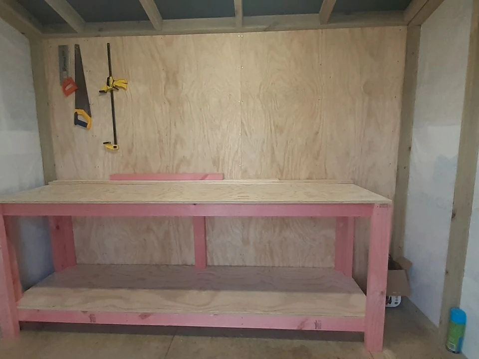 The workbench I built in the shed in 2021: a long ply-topped bench on a pink-painted timber frame, with a saw and clamps hanging on the wall behind